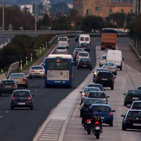 L'autopista de Llucmajor plena de vehicles (cotxes, motos, furgonetes, un autobús, un camió remolc, etc.) vistos des de darrera. De fons, una casa antiga mallorquina i un molí.