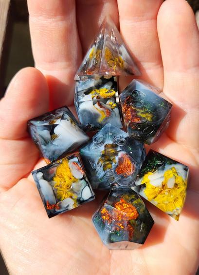 A TTRPG dice set in the palm of a hand. The dice contain yellow flowers, fiery orange metal foil and swirls of white and dark shimmery green.