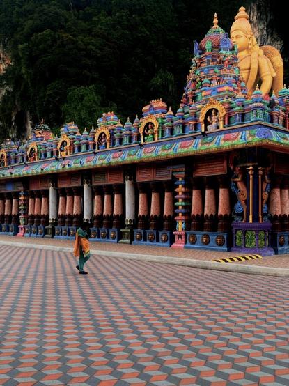 An image of a woman in a teal and orange sari walking across a temple in front of Batu Cave’s main statue of Murugan.