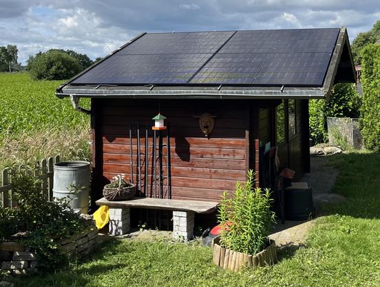 Foto: Eine schon etwas ältere Gartenhütte in dunkelbraun mit Satteldach. Die dem Betrachter zugewandte Dachseite ist mit vier PV-Panels der beschrienen Anlage belegt.