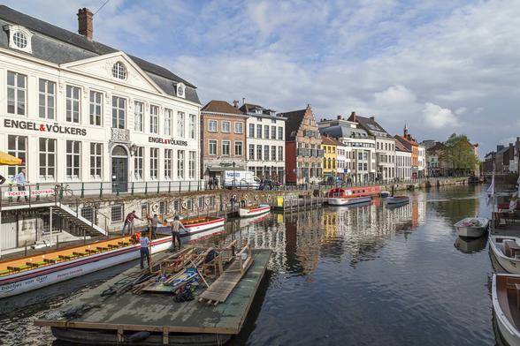 A bright, sunny, warm day with patchy white clouds in a deep blue sky overhead. We're standing on a low bridge over a wide canal running through the centre of a city with plenty of three-storey buildings along the water's edge, some of them clearly old and with a typically northern European style of stepped gable on the facade. Tour boats and narrow boats are tied up along both walls of the canal and those and the buildings reflect brightly on the rippled surface of the water. A wide, flat barge is in the foreground with some people on its littered deck apparently about to tie up next to another boat by a set of steps.