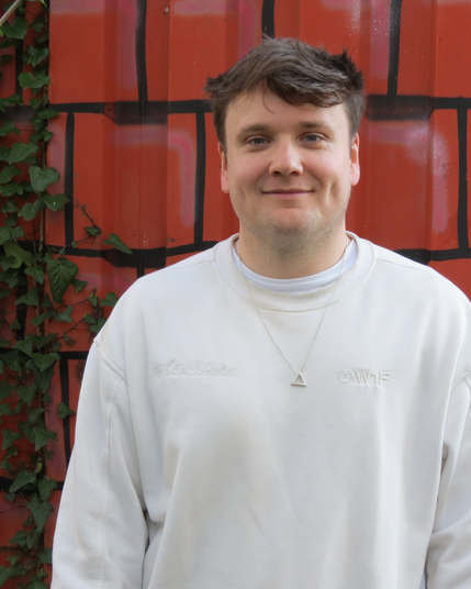 Patrik lächelt vor einer roten Steinmauer in die Kamera. Er trägt einen cremefarbenen Pullover mit einem kleinen, dreieckigen Anhänger vor einer rot-schwarz gemusterten Wand mit grünen Blättern.
