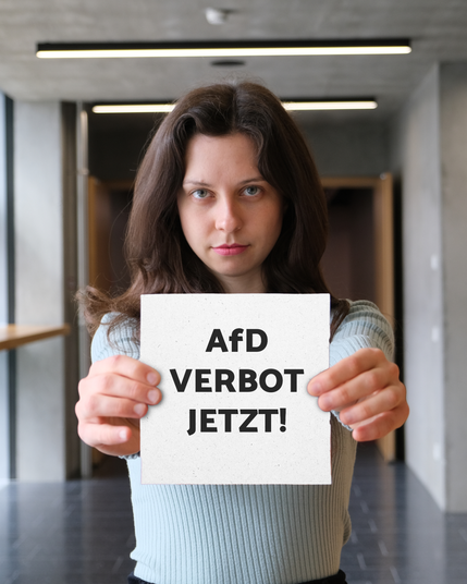 Zoe Mayer steht in einem modernen Flur und hält ein Schild direkt in die Kamera. Auf dem Schild steht in großer schwarzer Schrift: „AfD VERBOT JETZT!“. Sie blickt ernst und entschlossen.
