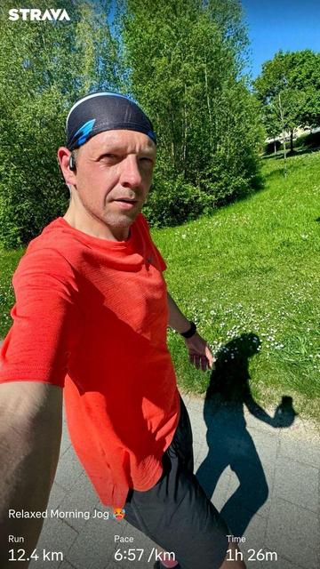 Me wearing a bright red shirt and a black bandana taking a selfie while jogging outdoors. The background features lush green trees and grass. The image displays jogging statistics, including distance (12.4 km), pace (6:57/km), and total time (1h 26m).