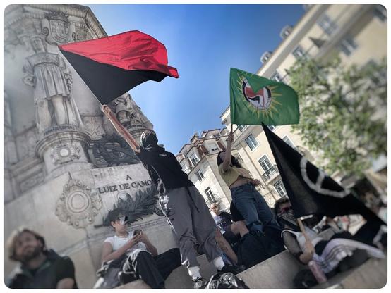 Eine Gruppe von Personen bei einem Protest oder einer Demonstration, die Flaggen mit verschiedenen Designs halten, darunter eine rote und schwarze Flagge, eine grüne Flagge mit einem Symbol und eine schwarze Flagge. Der Hintergrund zeigt ein dekoratives Denkmal mit Statuen und Gebäuden unter einem klaren blauen Himmel.