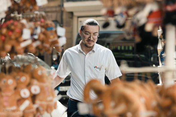 An Elis employee in a shirt pulls something that isn't really visible. The photo is taken through several portable displays of a shop selling Mannele plush toys.