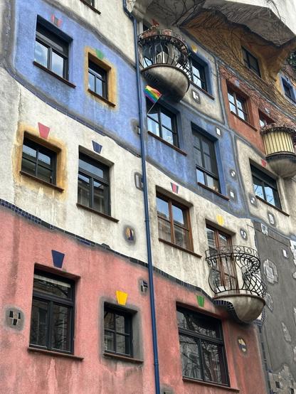 The exterior of a multi-storey apartment building, on which different sections are painted different colours, and delineated with wonky lines of ceramic tiles. There are windows, each a different size and shape than the others; around some of them, the wall within an arm’s reach is painted. There are a few small, semicircular balconies, some with ornate wrought-iron railings. One of the upper windows has a small LGBT flag flying from it.