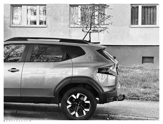 Schwarzweißfotografie: vor einem Wohnblock mit 3 sichtbaren großen Fenstereinheiten steht ein moderner kleiner Dacia SUV, dazwischen Rasen und ein kleiner Baum