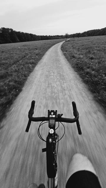 The image is a portrait-orientated black and white, first-person perspective shot from a bicycle on a gravel path. The path is visible in the foreground and recedes into the distance, curving slightly to the right. On either side of the path are grassy fields. In the background, there is a line of trees and a cloudy sky. Parts of the bicycle are visible in the foreground, including the handlebars, a cycling computer mounted on the handlebars, and a portion of the front wheel area with a bag attached. A knee or leg is also visible in the bottom right corner of the frame.