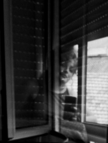 Fotografía en blanco y negro, borrosa, donde se ve a una mujer medio sonriente, reflejada en una ventana. Se forma un juego reflejos curioso.