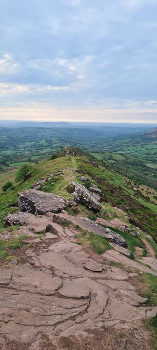 A narrow ridge, with a mix of rocky and grassy trail. Looking east, the background is the relative lowland and rolling hills of Herefordshire.
