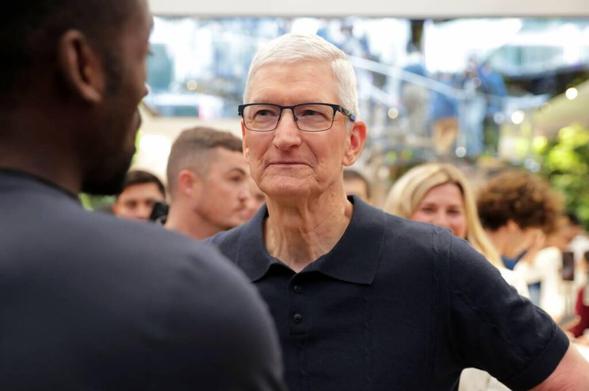 A group of people is gathered at a public or social event. The background shows a busy environment with several individuals, some engaged in conversation. The setting includes modern architecture and greenery, suggesting an indoor space with natural elements. In the foreground, Apple CEO Tim Cook, wearing a dark polo shirt and glasses, is engaged in conversation with another individual. The image captures a moment of interaction and social engagement.