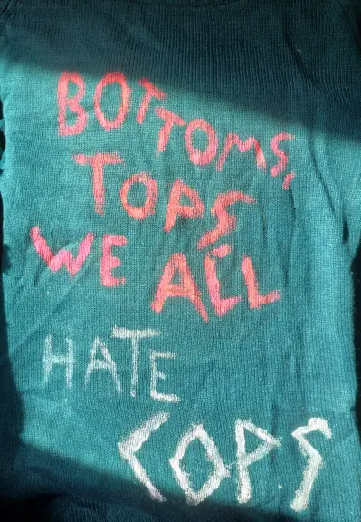 A green/turqoise sweater, 'Bottoms, Tops, We All Hate Cops' painted raggedly on it.
