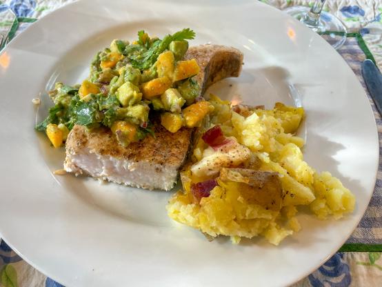 Piece of grilled fish covered with gooey mound of diced yellow mango, diced avocado, and cilantro. Nearby is a pile of potatoes studded with melted cheese and bacon.