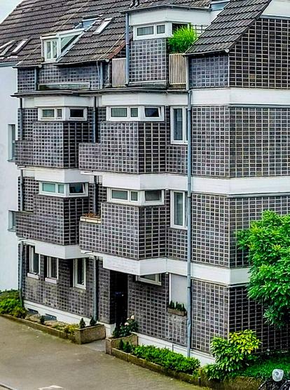 Das Bild zeigt die Fassade eines mehrstöckigen Wohngebäudes in einem modernen, funktionalistischen Stil. Die Fassade ist geprägt durch eine regelmäßige Rasterstruktur aus dunklen, kleinformatigen Elementen, die wie Klinker oder Fliesen wirken, und hellen, vermutlich verputzten oder betonierten horizontalen Bändern, die die Geschosse optisch trennen.
Jede Wohneinheit verfügt über einen oder mehrere rechteckige Balkone, die in die Fassadenstruktur integriert sind. Die Balkonbrüstungen bestehen aus demselben dunklen Rasterelement wie die übrige Fassade. Die Fenster sind rechteckig und scheinen über eine moderne Verglasung zu verfügen.
Das Gebäude hat ein schräges Ziegeldach mit mehreren Dachgauben. Im obersten Geschoss sind einige Balkone etwas zurückgesetzt und bieten Platz für kleine Grünpflanzen.
Im Erdgeschoss sind Hauseingänge und kleine Vorgärten mit niedriger Bepflanzung zu erkennen. Vor dem Gebäude verläuft eine asphaltierte Straße.