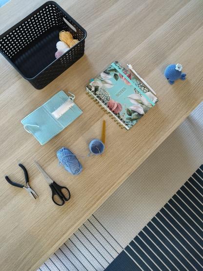 Picture of a part of a desk and the floor with a blue carpet. One the desk is a crochet project in progress with blue yarn, scissors, needles, a notebook, a pen, a black basket with some yarn in different colors, and a whale amigurumi.