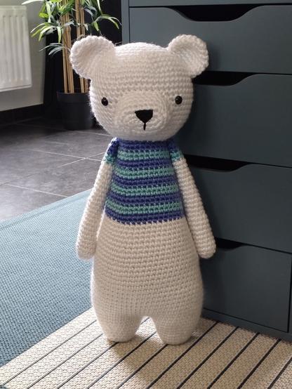 Picture of a crocheted polar bear standing upright leaning against some drawers of a desk. it's wearing a blue-striped shirt.