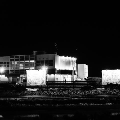 This black and white nighttime photograph captures a group of illuminated commercial buildings in Kushiro. The structures feature a modern architectural style with glass panels and rectangular forms. Several bright advertising banners or billboards are visible in front of the buildings, glowing white against the pitch-black night sky. Spotlights mounted on the buildings create starbursts of light. A railing or fence runs along what appears to be a railway track in the foreground, with snow or cleared ground visible. The stark contrast between the bright, lit structures and the surrounding darkness creates a dramatic nighttime urban landscape.