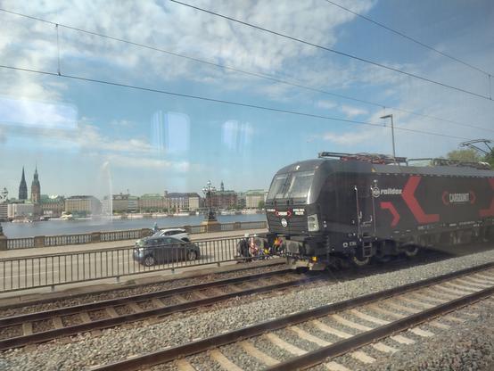Photo taken from a train going over a bridge in front of the Hamburg Binnenalster waterfront. The train is overtaking a black and red freight locomotive, and in the background you can see famous Hamburg landmarks.