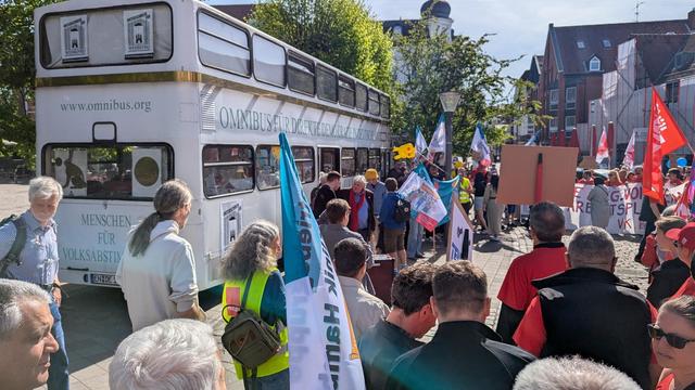 Der Omnibus (weißer Doppeldecker, Aufschriften: www.omnibus.org / Menschen für Volksabstimmungen, Omnibus für direkte Demokratie), daneben eine Menschenmenge: Leute vom Omnibus und eine Mai-Demo mit Plakaten und Fahnen