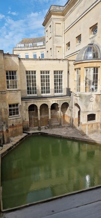 Picture showing the roman baths. An historic pool filled with green water. Sandstone buildings with large windows. Bright blue sky with some clouds