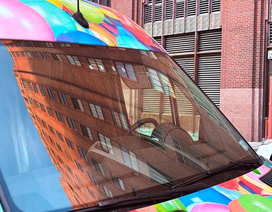 A van parked in Tribeca. The windshield is reflecting a large brick building and a blue sky. The van is decorated by a balloon themed paint job