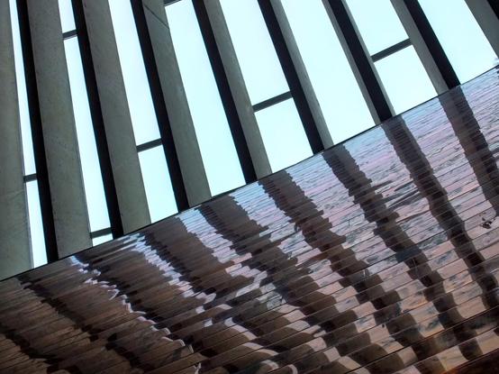 This image showcases an architectural detail, viewed from below. It features a series of vertical slats or beams that create a pattern against a light sky. The surface below appears reflective, capturing the lines and shapes overhead, adding an abstract quality to the composition. The interplay of light and shadow enhances the visual interest.