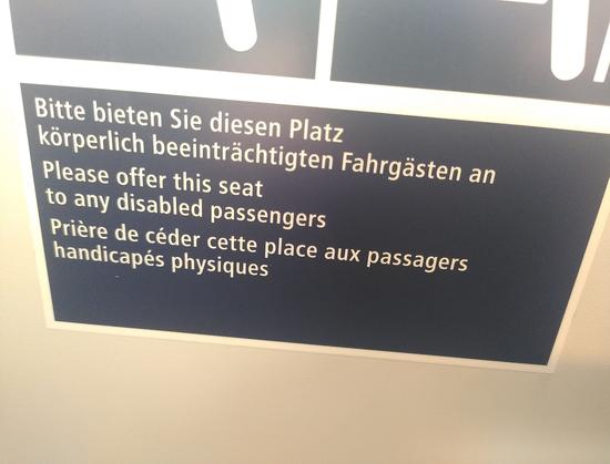 Ein mehrsprachiges Hinweisschild für Sitzplätze mit mehr Beinfreiheit. Im Text wurde "körperlich beeinträchtigt" mit "disabled" übersetzt (oder andersrum)