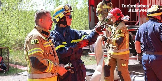 Eine Person in Einsatzkleidung des Technischen Hilfswerks spricht mit einer Person in Feuerwehruniform mit dem Aufdruck PSP. Im Hintergrund rollt eine weitere Person in Feuerwehruniform einen Schlauch auf. Weiterhin befinden sich im Hintergrund ein grüner Wald und ein großes rotes Fahrzeug sowie zwei weitere Personen in Kleidung des Technischen Hilfswerks und der Feuerwehr.
