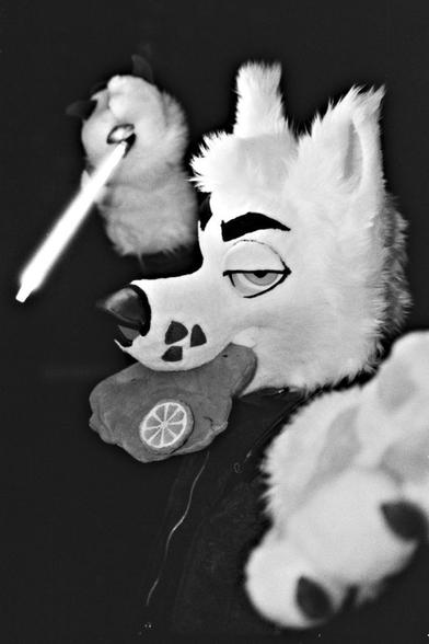A black and white photo of a canine fursuiter. He looks into the camera with a large Schnitzel in his muzzle. He holds one paw to the camera, the other one holds a light-saber.