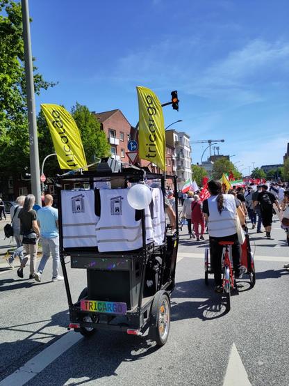 Ein großes dreirädriges Bike mit Tricargo-Flaggen und buntem Tricargo-Nummernschild. An den Lautsprecherboxen hängen HWF-Westen. Erik radelt daneben auf einem Lastenrad. Es laufen in der strahlenden Sonne viele Menschen in die gleiche Richtung