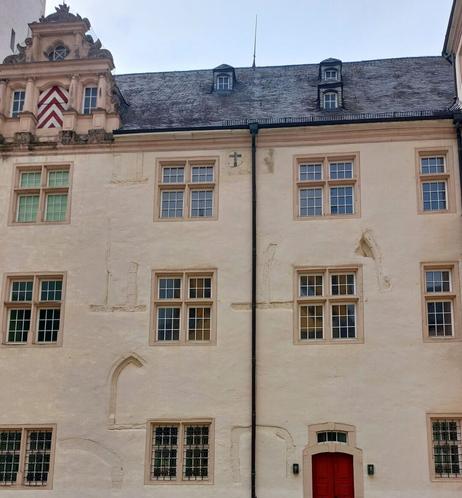 Foto einer beige gestrichenen Fassade eines dreistöckigen Gebäudes mit zusätzlichen Geschossen im Dach.
An der Fassade sind regelmäßig rechteckige Fenster mit jeweils 2 geteilten Fensterflügeln angeordnet. Dazwischen sind im Mauerwerk nicht vollständig verputzt die Umrisse älterer gemauerter Fensterrahmen auf unterschiedlichen Höhe  zu sehenn mit unterschiedlichen Höhen und verschiedenen Bogenformen.