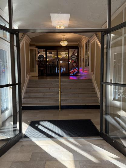 Ornate entry hallway with marble steps leading to a pinball parlor. Neon "Open" sign and warm chandelier create an inviting, nostalgic atmosphere.