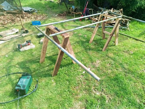 A lawn with junk and tools spread around. A section of fence is opened up in the background. In the foreground, a half finished gate with freshly extended legs is clamped to two sawhorses standing in the grass. On the floor is a small green fluxcore welder, a battery angle grinder and some other stuff. A wire brush is on the saw horse as well.

The red farm jack rests in the background, having extracted the old gate post.