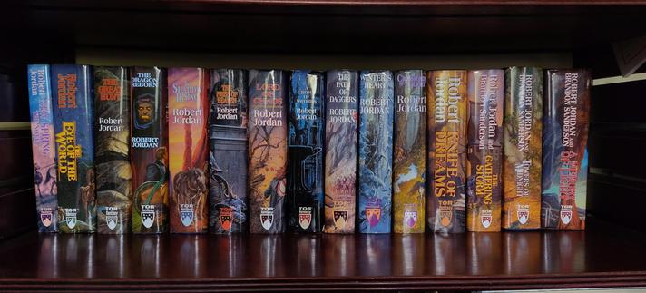 a wide angle view of a large, dark wood bookshelf. On it are 15 books, all of them quite thick, with colourful artwork on the covers. It's the entire Wheel of Time series of books, plus New Spring novel, in chronological order