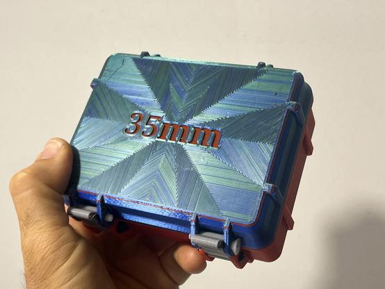 A closed 3d printer box with an embossed “35mm” on the top. A cool pattern is created by the multicolored filament in a radiating pattern.