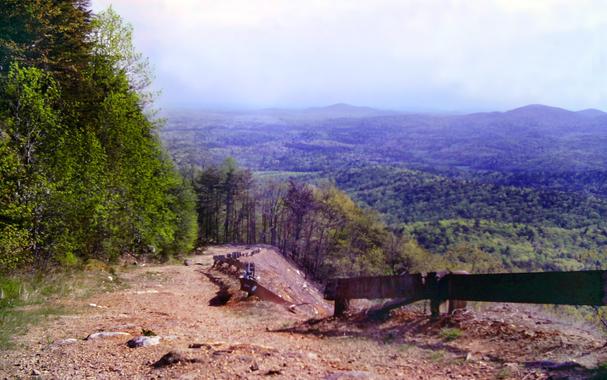 Standing high up the steep slope of a mountain looking down at forested foothills and valleys far below. The sky is hazy and whitewashed, but bright and sunny. The forest down below is very green. Leaves are just starting to come in up here and are still small. We are climbing a rocky dirt fire road up this steep slope. There are wooden guardrails, broken in a couple of places, protecting any drivers who use this road from a very steep drop to our right. Like the road, that slope is also naked reddish clay soil and rocks. The forest presses up against the left side of the road. Some distance below us the steep drop-off on our right eases slightly and there are trees on that side as well.