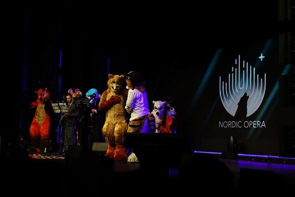 Photo of an indoor show stage, with some furry characters in the middle of performing a live opera. To our left are the musicians, including Vozzy the Fox currently playing the violin. At the centre of the stage, Chester the Geroo and Dan the Singing Tiger are currently engaged in a back-and-forth duet. To our right, the Nordic Opera logo is projected on the wall in the background. The stage is dark and dimly lit, with a spotlight on the two conversational singers.