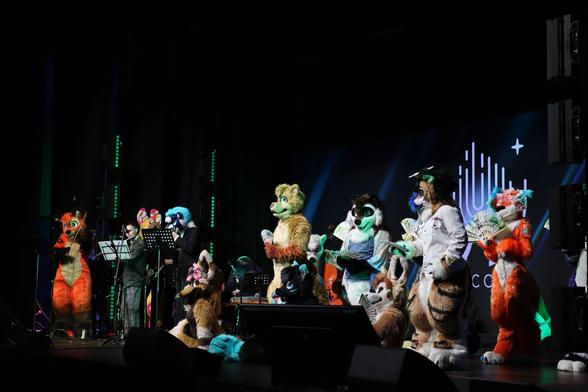 Photo of an indoor show stage, with a full cast of furry characters in the middle of performing a live opera. To our left are the musicians, including a bright orange fox currently playing the violin, while the centre and right is filled with various fuzzy creatures sitting and standing around, posing or singing. Among them are a brown cross fox, a tanuki and a tiger wearing a naval uniform hat and shirt. Most of them are holding traditional Japanese folding fans. The stage is dark, but all the characters are brightly lit by white and green stage lights for this particular scene.