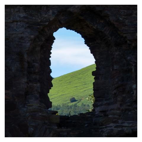 View of a lush green hillside and clear blue sky framed by an old, weathered stone archway, creating a natural window to the vibrant landscape beyond.