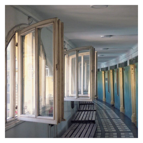 This is a square format colour image of the (empty!) changing rooms of the Szechenyi Baths in Budapest. Open windows dominate the foreground and allow light to fall from left to right through the frame. The closed doors of the changing cubicles curve away from the right. Ornate tiles cover the floor and wooden benches sit underneath the open windows. The predominant colours are pale blues and yellows.