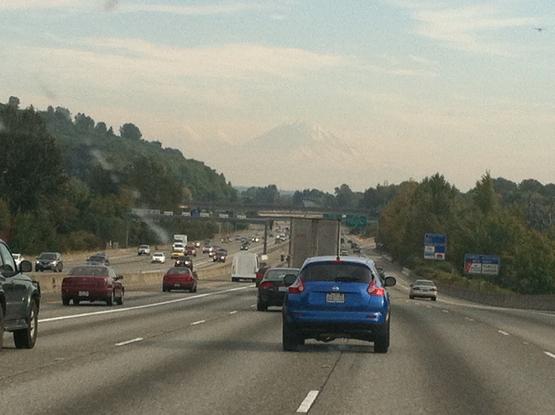 A five-lane-wide interstate with a handful of cars and a snow-capped mountain ahead nearly hidden by the smoke in the air.