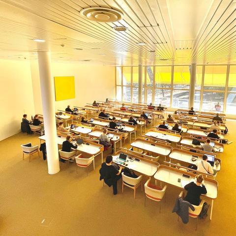 A spacious, brightly lit study room with rows of desks, filled with students working on laptops. Large windows bring in natural light.