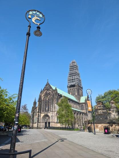 A medieval cathedral across a cobbled square under a bright blue sky. In the foreground on the left is a decorative lamp post displaying a fish, a bell, a tree, and a bird