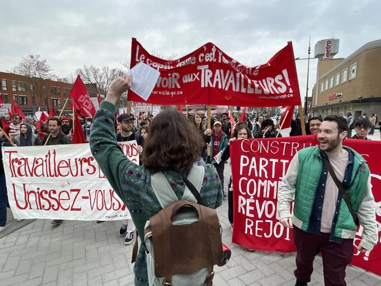 During a rally for the International Worker's day, people are holding «Workers unite!» banners while a young man, seen from behind, shouts slogans.