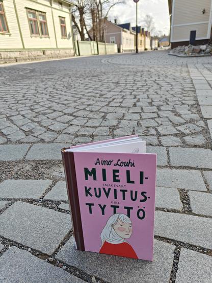 Kuvassa Mielikuvitustyttö-kirja kuvattuna Vanhan Rauman kadulla. Takana näkyy vanhoja puutaloja.