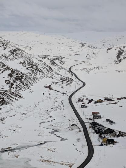 Black road winding in a snowy landscape. A few houses there and there. The sky is as white as the snow.