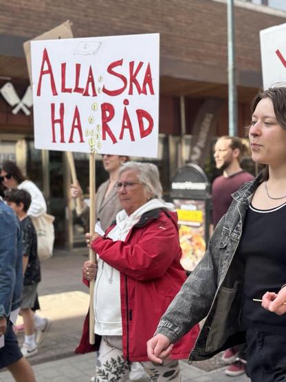 Människor i tåg i Skövde. En kvinna i röd jacka håller upp en skyld som säger "Alla ska ha råd".