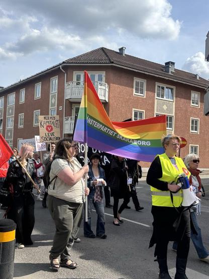 Manniskor på gatan som håller i banderoller och skyltar. Framst en banderol i regnbågfärg som säger "Vänsterpartiet Skaraborg".