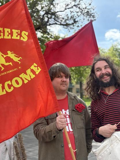 Two människor med röda flagor, en med teksten 'refugees welcome'.